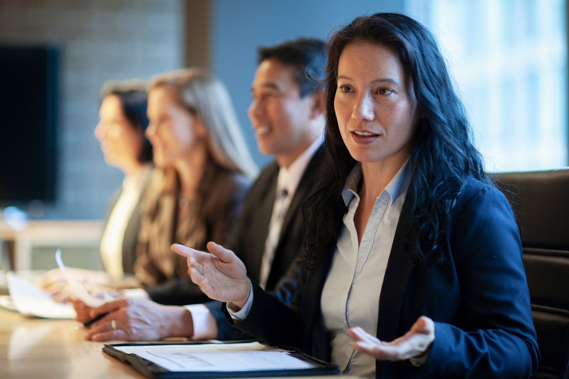 Multiracial Caucasian Thai ethnicity mature woman businesswoman explaining talking with multiracial group of work colleagues in business meeting talking discussing plans ideas in businesswear in board room conference table
