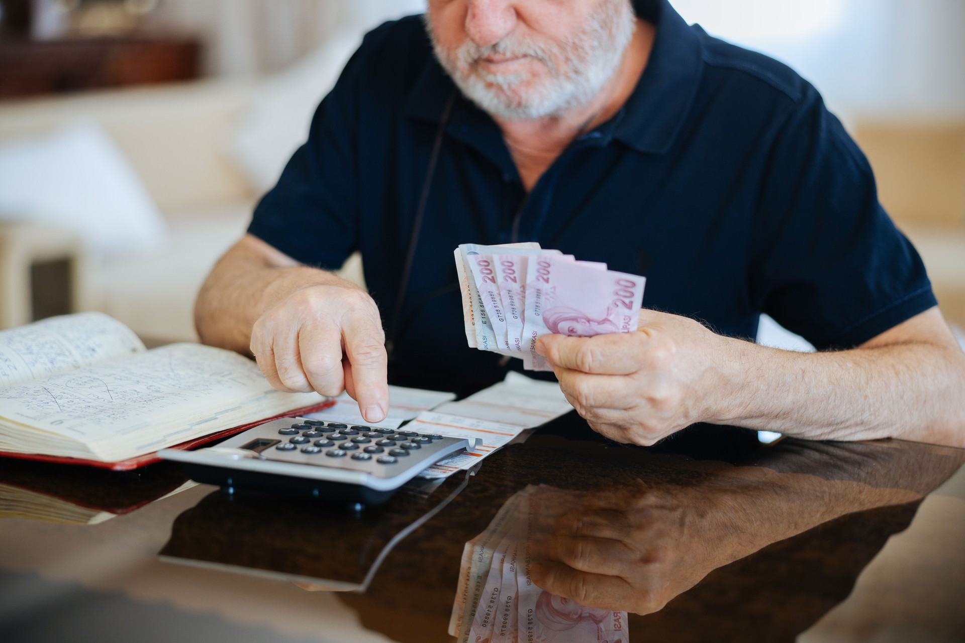 Anxious senior man managing finances, holding cash and using calculator at home